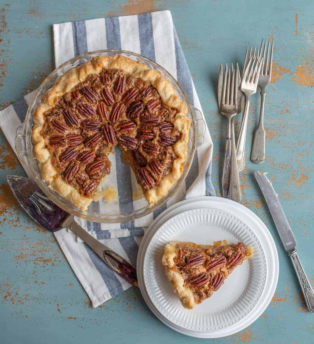An overhead shot of a pecan pie in a clear dish on top of a folded blue and white striped dish towel. On the lower left hand side of the pie is a silver pie cutter. To the right of the pie are varying sizes of silver forks. Under the forks is a slice of the pecan pie that is on a fancy white plate. Under that plate are varying sizes of plain white dishes. On the right side of the slice of pie is a silver butter knife. These are all resting on a turquoise painted table that has wood showing through in spots.