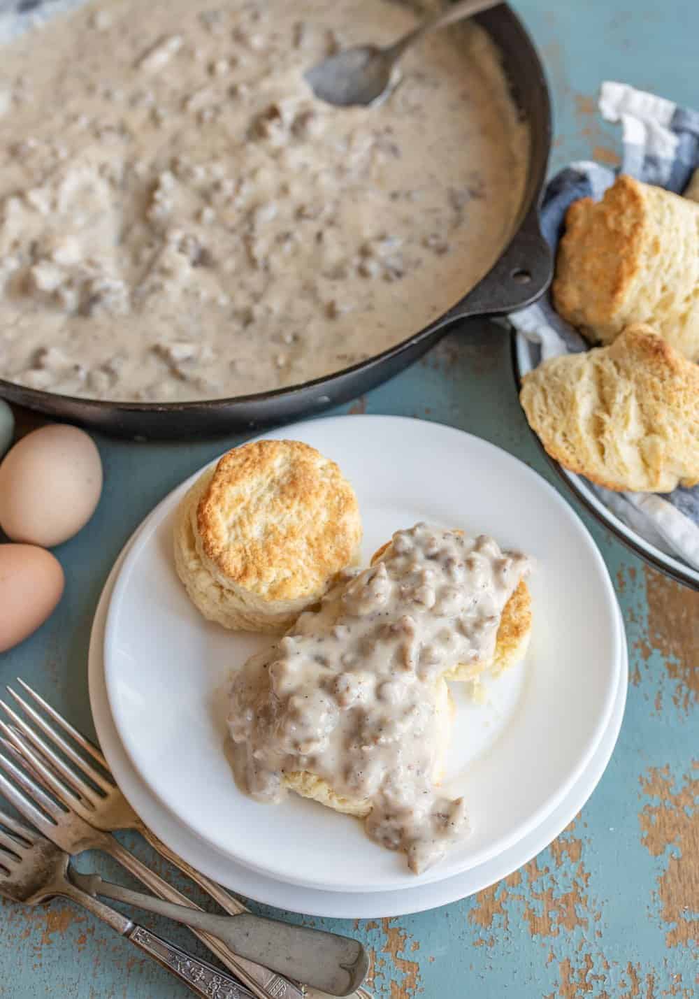 White plate with biscuit on it and a biscuit topped with gravy on a light blue table with a pan of gravy in the background