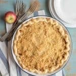 A Dutch apple pie recipe takes center stage on a striped cloth, surrounded by forks, an apple, and stacked white plates. The crumb-topped delight is set against a rustic, blue-painted background.