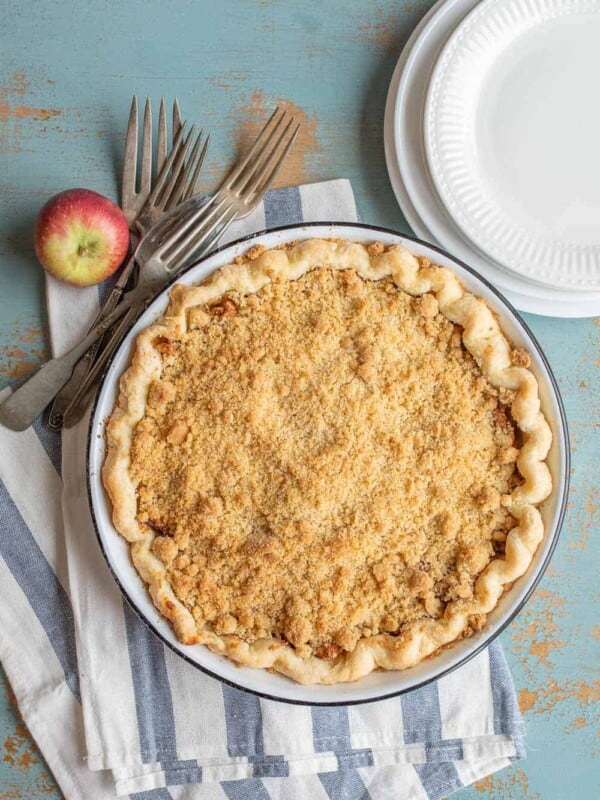A Dutch apple pie recipe takes center stage on a striped cloth, surrounded by forks, an apple, and stacked white plates. The crumb-topped delight is set against a rustic, blue-painted background.