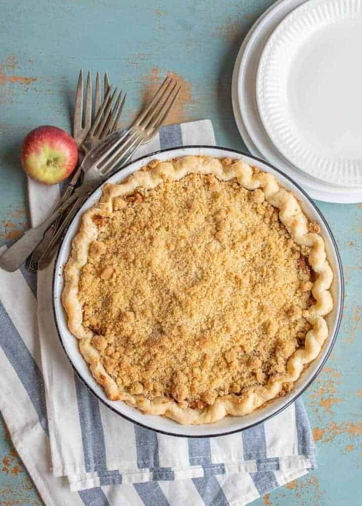 A Dutch apple pie recipe takes center stage on a striped cloth, surrounded by forks, an apple, and stacked white plates. The crumb-topped delight is set against a rustic, blue-painted background.
