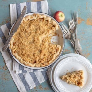 A crumb-topped pie from a dutch apple pie recipe sits in a pan with one slice missing. The missing slice rests on a white plate nearby. An apple, forks, and a knife are arranged on a striped cloth atop a rustic blue surface, inviting you to try your hand at this classic recipe.