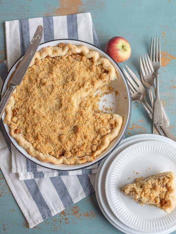 A crumb-topped pie from a dutch apple pie recipe sits in a pan with one slice missing. The missing slice rests on a white plate nearby. An apple, forks, and a knife are arranged on a striped cloth atop a rustic blue surface, inviting you to try your hand at this classic recipe.