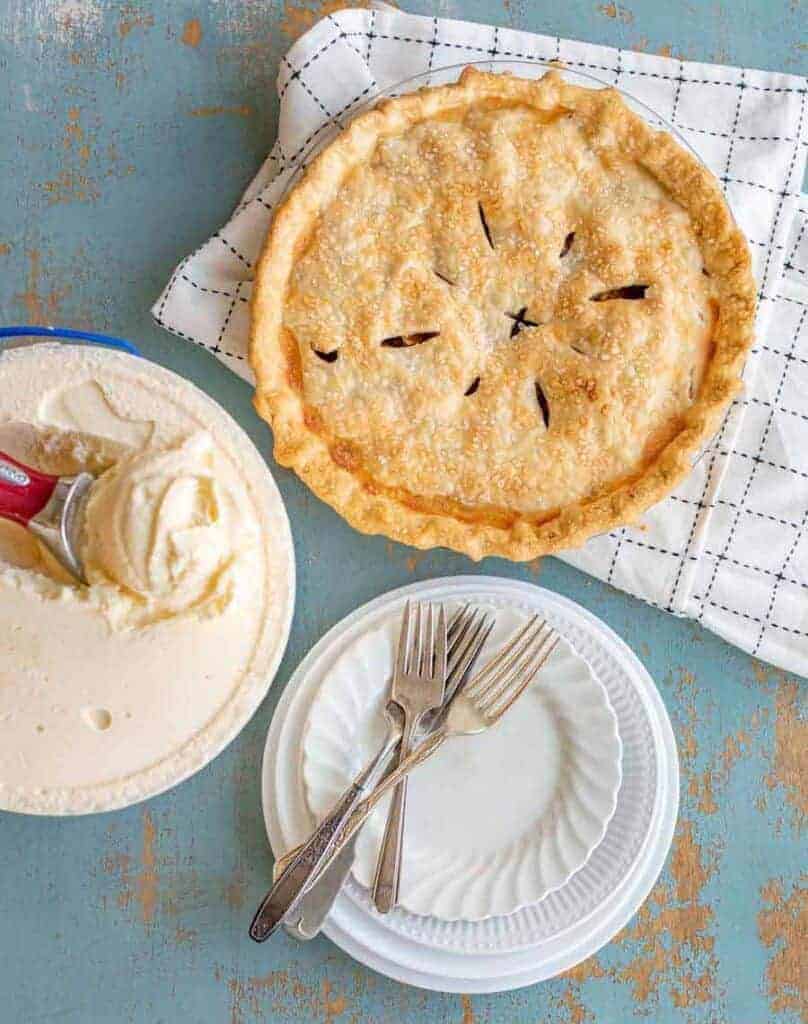 whole apple pie next to plates and a gallon of ice cream ready to be served