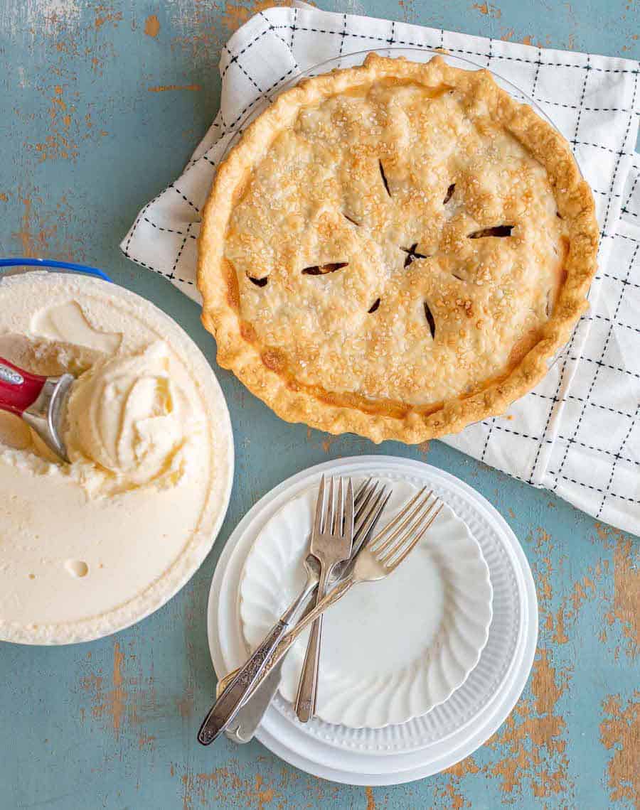 whole apple pie next to plates and a gallon of ice cream ready to be served