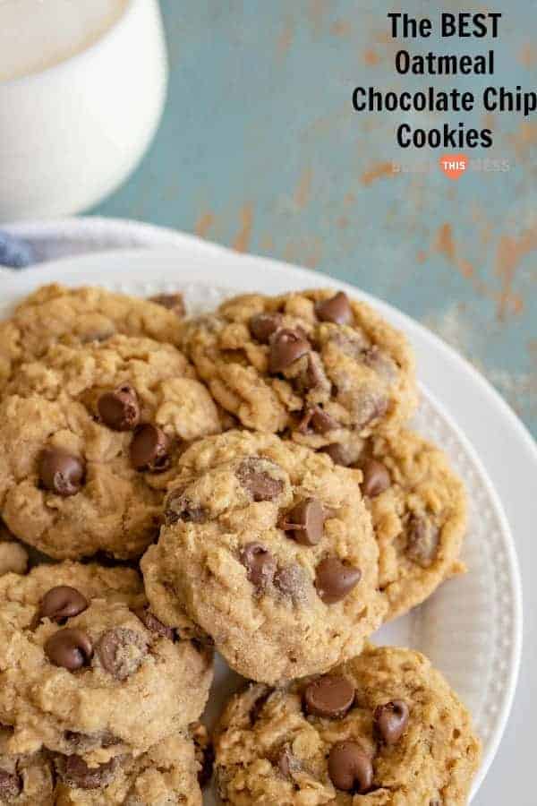 A plate of the best oatmeal chocolate chip cookies is stacked in the foreground. The words The BEST Oatmeal Chocolate Chip Cookies are written in the upper right corner. A partial view of a cup is visible in the background.