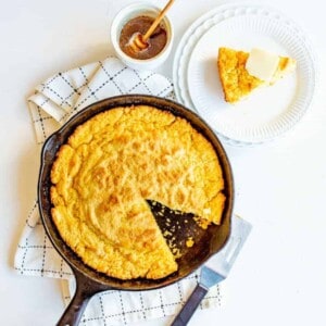 A skillet cornbread sits golden on a checkered towel, a slice missing. Nearby, a plate holds a buttery serving of skillet cornbread, ready to be drizzled with honey from the dish and honey dipper alongside.