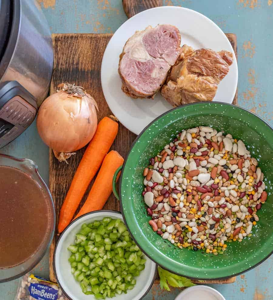 green bowl of dried beans with white bowl filled with diced celery on cutting board with whole carrots yellow onion and ham