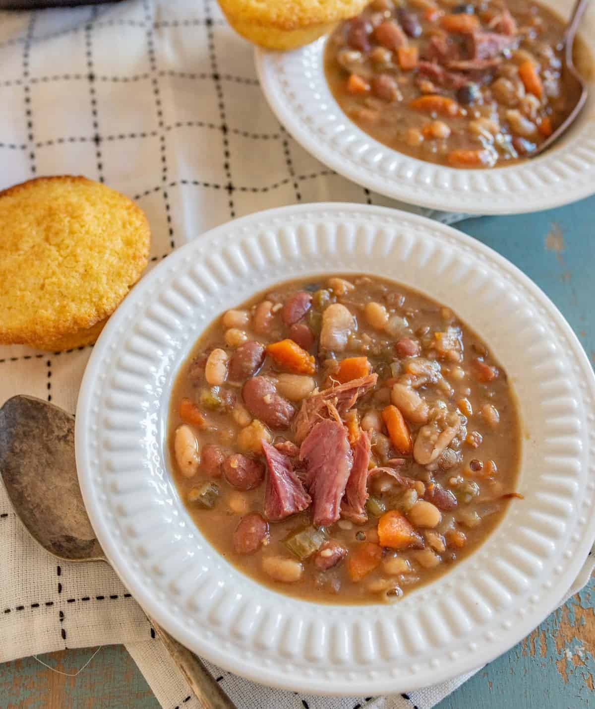 two white bowls of bean 15 bean soup with a corn muffin on white towel with a spoon 
