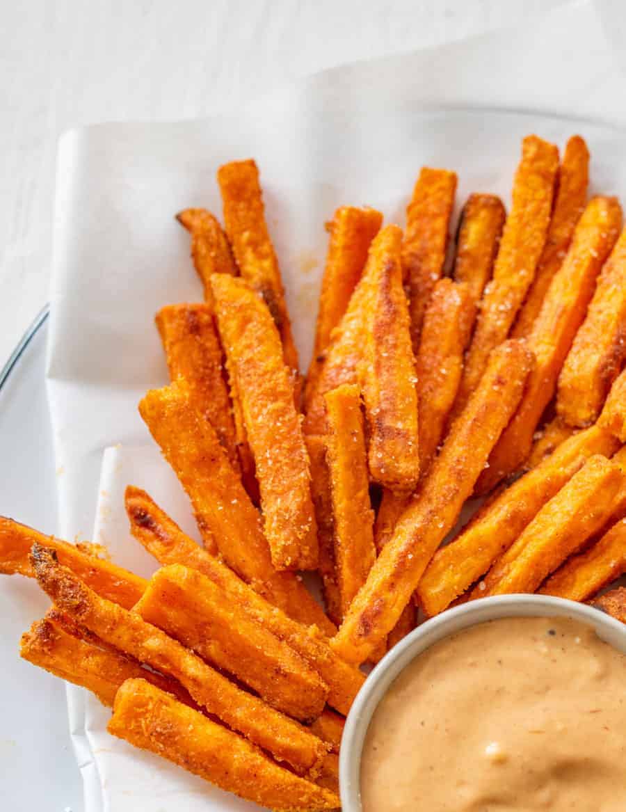 A close-up of a pile of baked sweet potato fries on white parchment paper on a white plate with a small bowl of light red dipping sauce. 