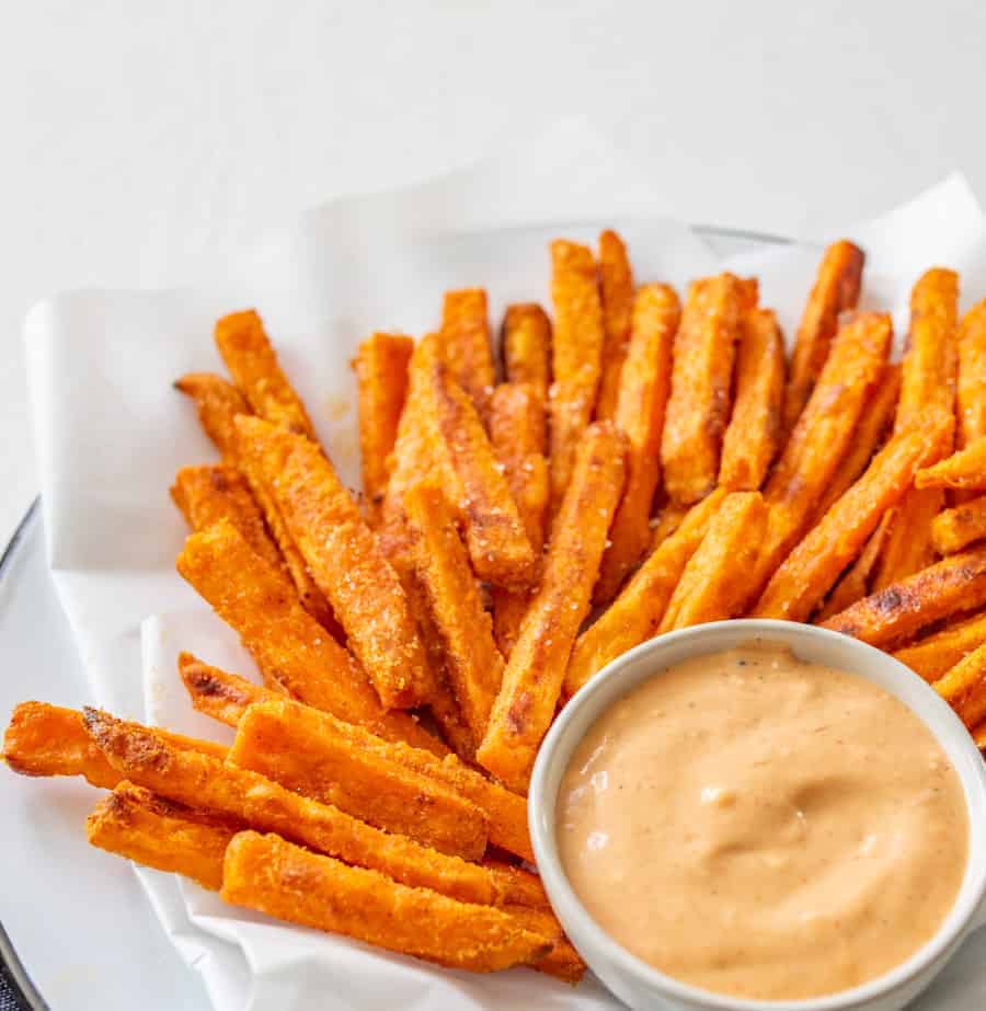 Image of a pile of sweet potato fries on a piece of white parchment paper on a white plate with a small bowl with a light red dipping sauce. 