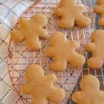 Gluten free gingerbread cookies shaped like people cool on a round copper wire rack. They rest on a white cloth with a black grid pattern. The unadorned cookies are evenly spaced, inviting the warmth of the season into your home.