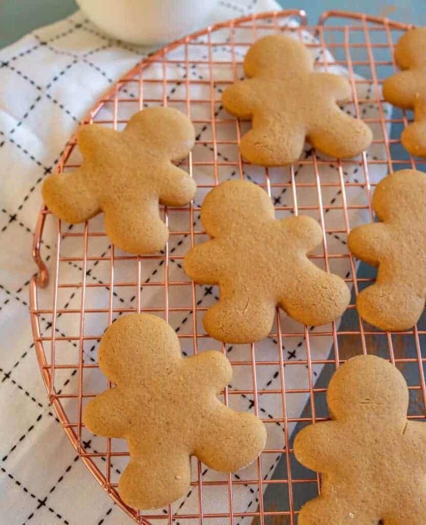 Gluten free gingerbread cookies shaped like people cool on a round copper wire rack. They rest on a white cloth with a black grid pattern. The unadorned cookies are evenly spaced, inviting the warmth of the season into your home.