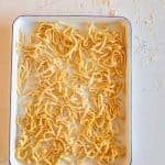 A white rectangular tray holds uncooked, homemade noodles, their yellowish strands scattered randomly. The background features a light-colored surface dusted with flour, enhancing the rustic charm of this homemade delight.