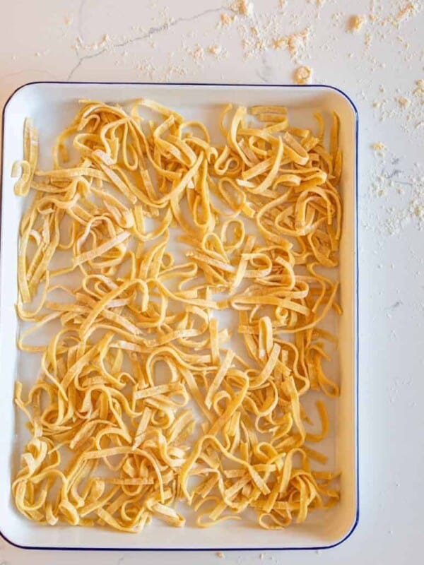 A white rectangular tray holds uncooked, homemade noodles, their yellowish strands scattered randomly. The background features a light-colored surface dusted with flour, enhancing the rustic charm of this homemade delight.