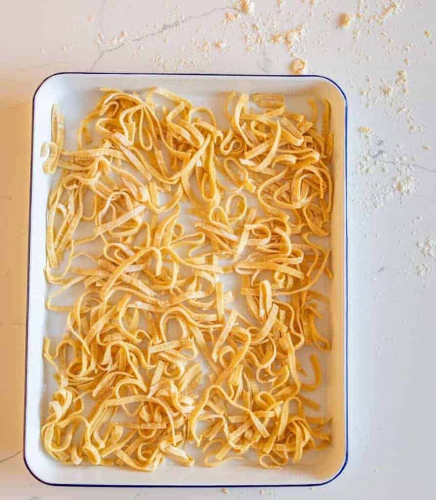 A white rectangular tray holds uncooked, homemade noodles, their yellowish strands scattered randomly. The background features a light-colored surface dusted with flour, enhancing the rustic charm of this homemade delight.
