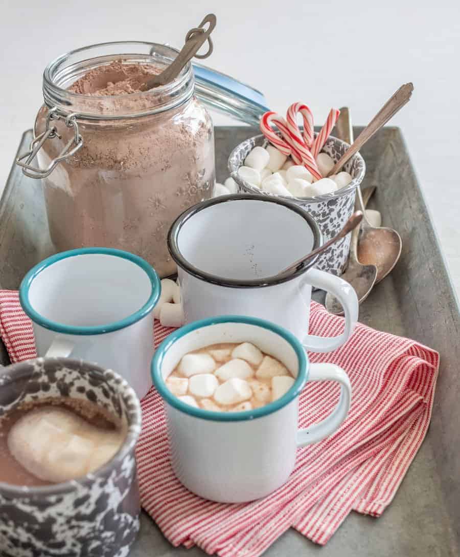 Image of a jar clear glass jar filled with homemade hot cocoa mix, a white enamel cup with a blue rim filled with hot cocoa, and a white enamel cup with grey swirls with mini marshmallows and small candy canes, a red and white striped cloth napkin, and a couple small spoons on a metal tray.