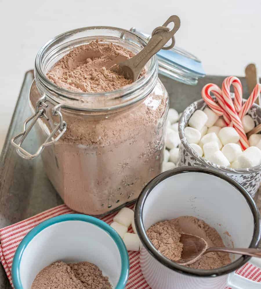 Image of a jar clear glass jar filled with homemade hot cocoa mix, two enamel cups with a couple tablespoons of hot cocoa mix in each, and a white enamel cup with grey swirls filled with mini marshmallows and small candy canes, a red and white striped cloth napkin, and a couple small spoons on a metal tray.