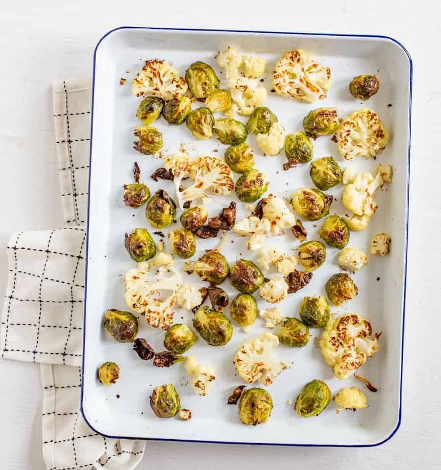 Image of roasted Brussels sprouts and cauliflower on a white enamelware baking sheet resting on a white napkin with black accents. 