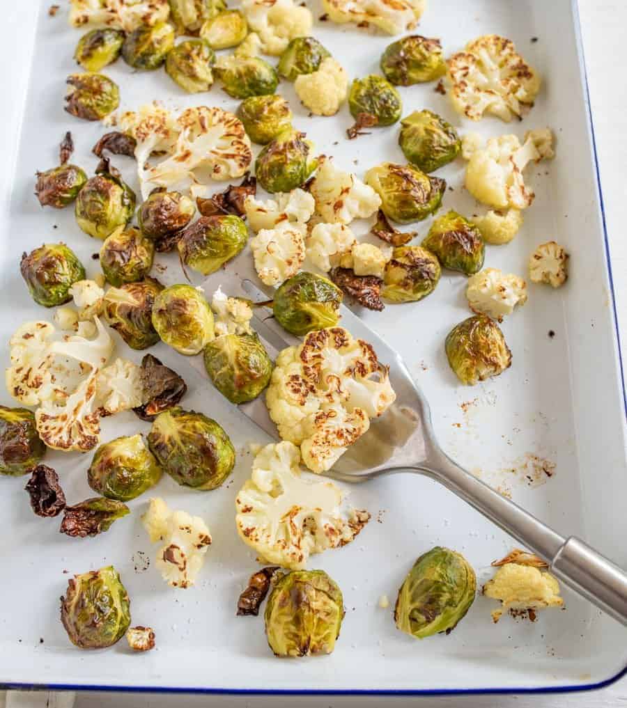 Image of roasted Brussels sprouts and cauliflower on a white enamelware baking sheet with a blue rim and a fork. 