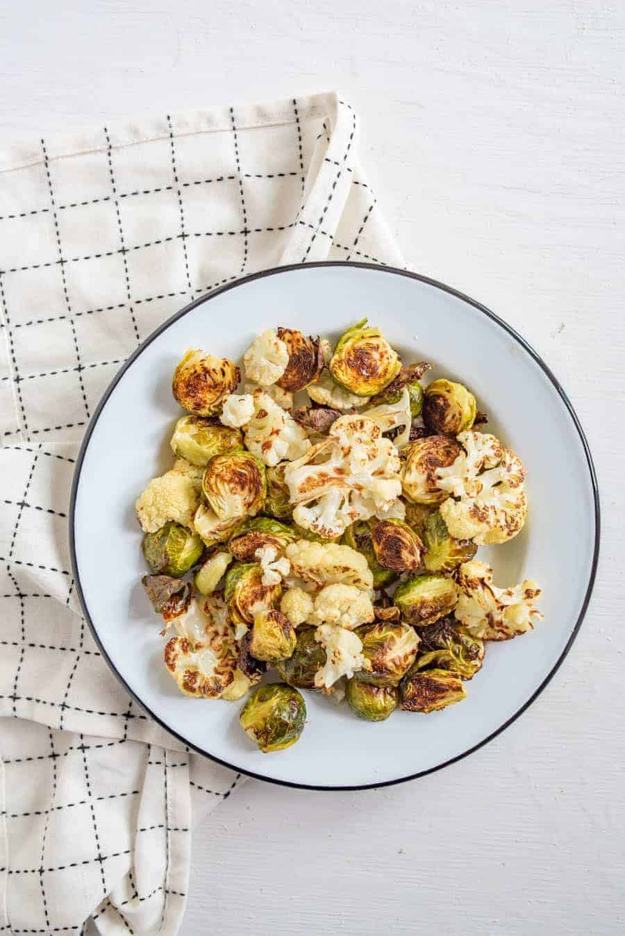 Image of roasted Brussels sprouts and cauliflower on a white plate with a black rim on a white napkin with black accents. 