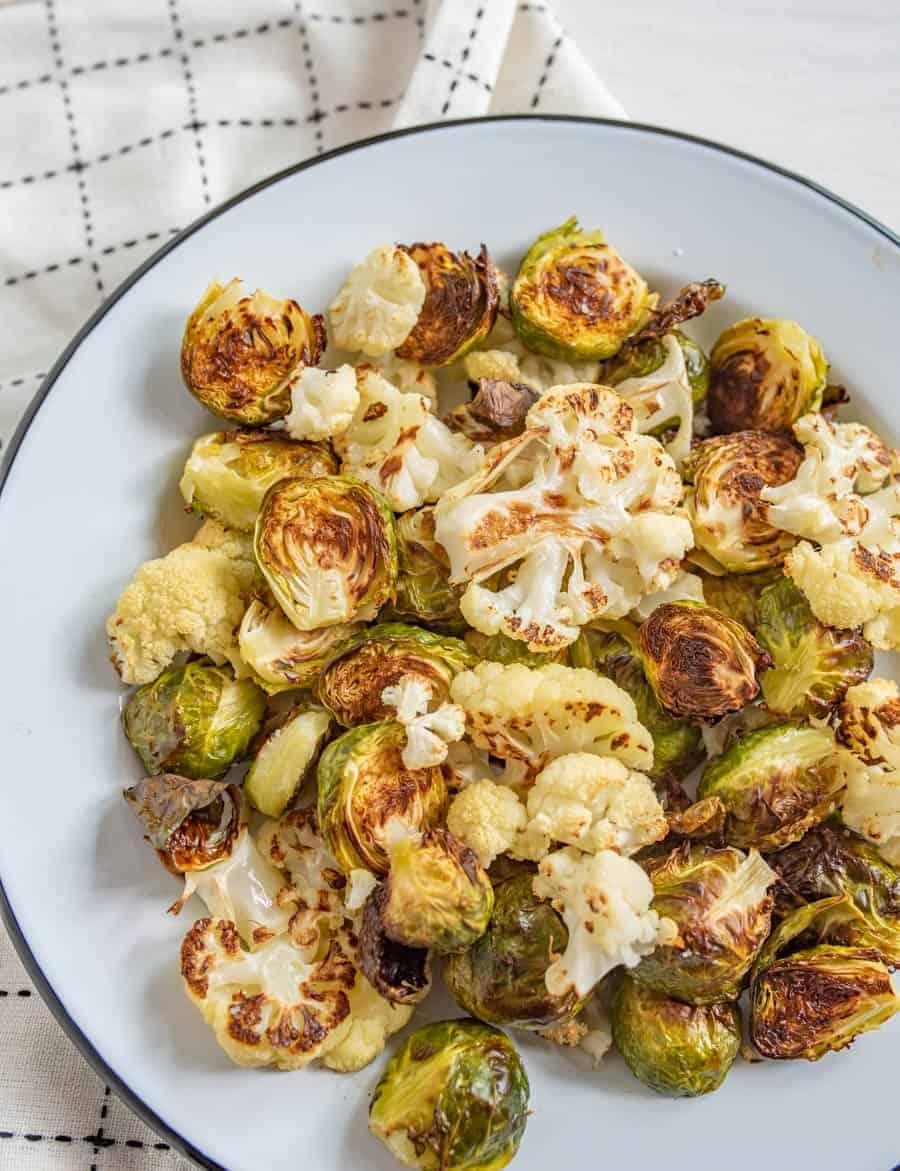 Image of roasted Brussels sprouts and cauliflower on a white plate with a black rim on a white napkin with black accents. 