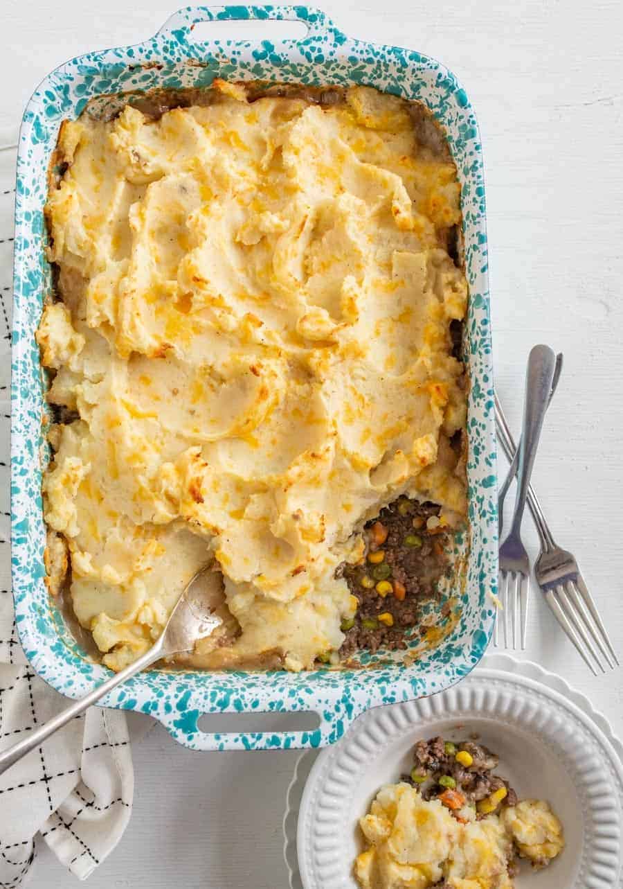 A serving of shepherd's pie in a white bowl with three forks and a white and blue speckled enamel pan with the rest of the pie in the background. 