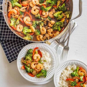 A skillet of shrimp stir fry recipe with broccoli, red peppers, and mushrooms sits beside two plates of white rice topped with the savory dish. Four forks rest on a checkered cloth nearby.