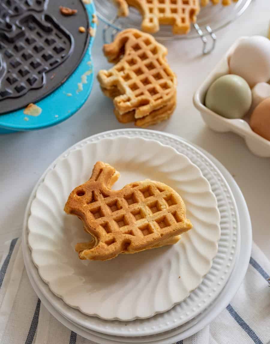 Image of a whole wheat waffle in the shape of a rabbit on a white plate on top of a white napkin with blue stripes. 