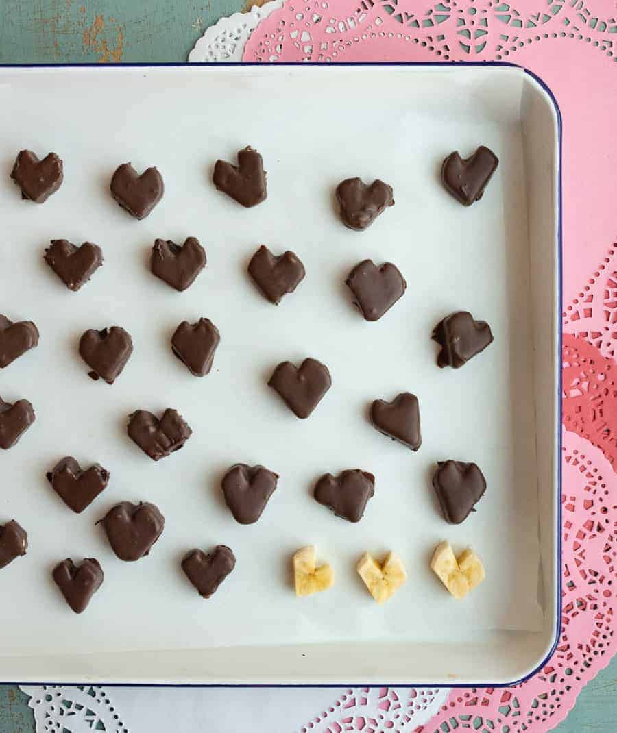 Slices of banana dipped in chocolate on a cookie sheet with parchment paper with red, pink, and white paper doilies in the background. 