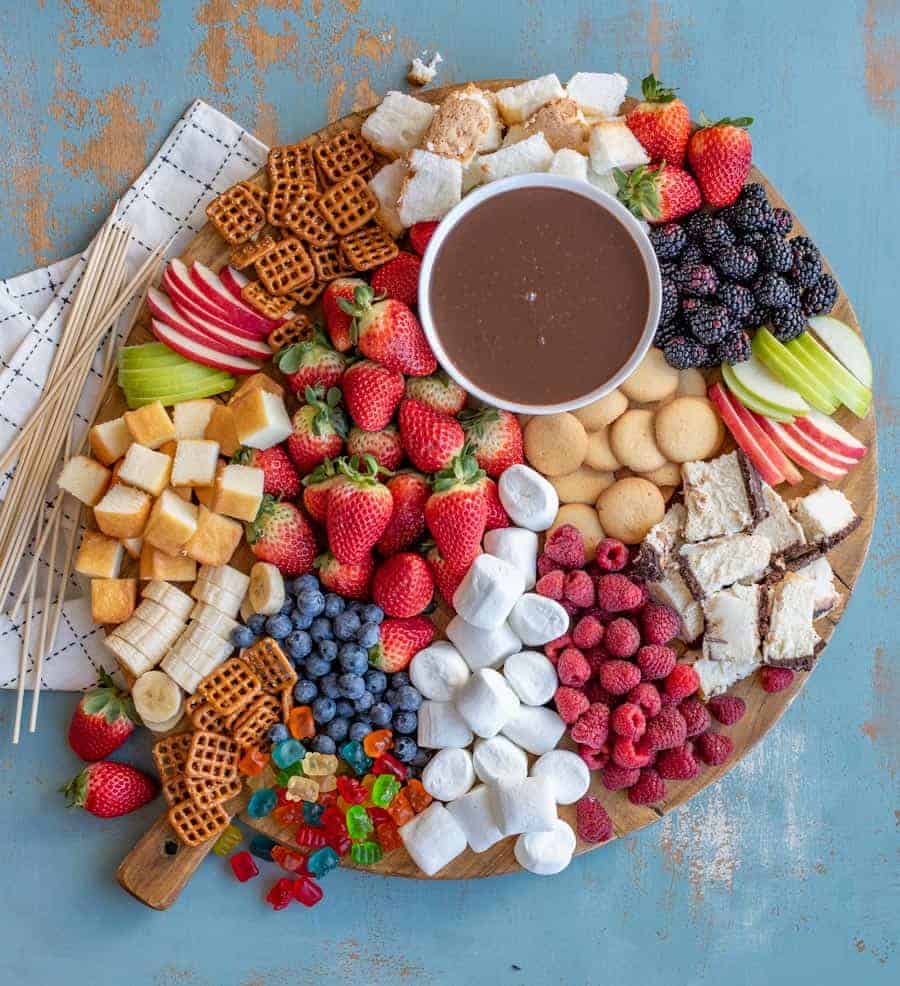 A board with a bowl of chocolate fondue surrounded by things to dip in it, including strawberries, vanilla wafers, apple slices, blackberries, pound cake, raspberries, and angel food cake. 