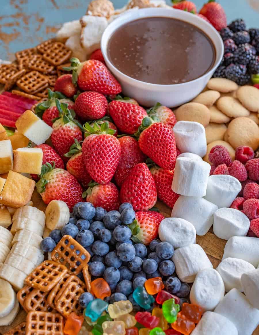 A board with a bowl of chocolate fondue surrounded by things to dip in it, including strawberries, pretzels, vanilla wafers, apple slices, blackberries, pound cake, raspberries, and angel food cake. 