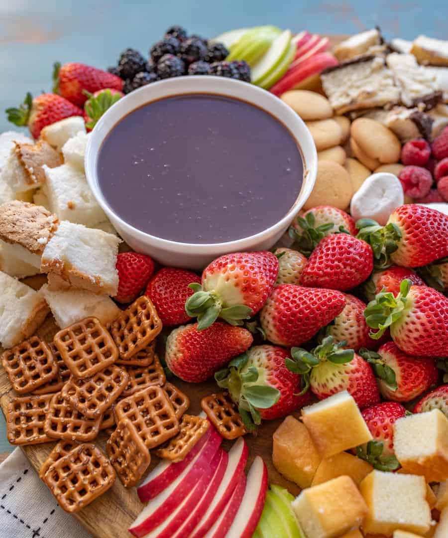 A board with a bowl of chocolate fondue surrounded by things to dip in it, including strawberries, pretzels, vanilla wafers, apple slices, blackberries, pound cake, raspberries, and angel food cake. 