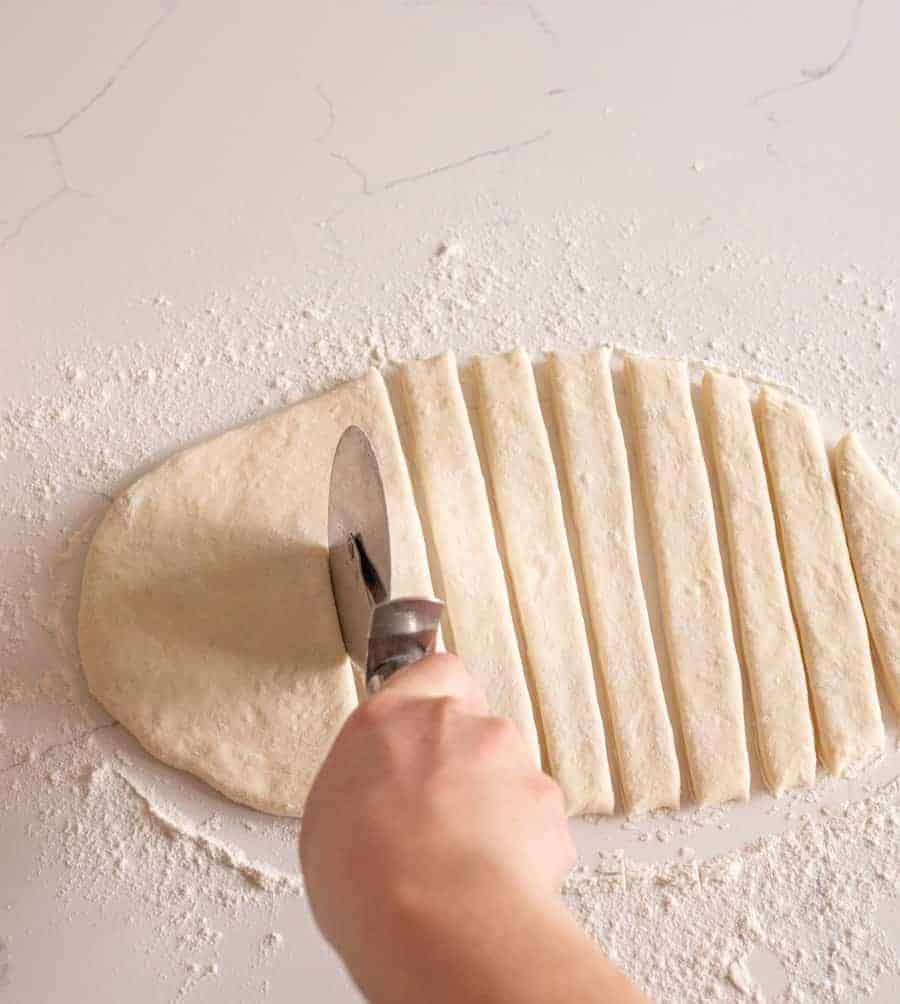 a pizza cutter being used to slice a sheet of breadstick dough into long strips on a floured counter.