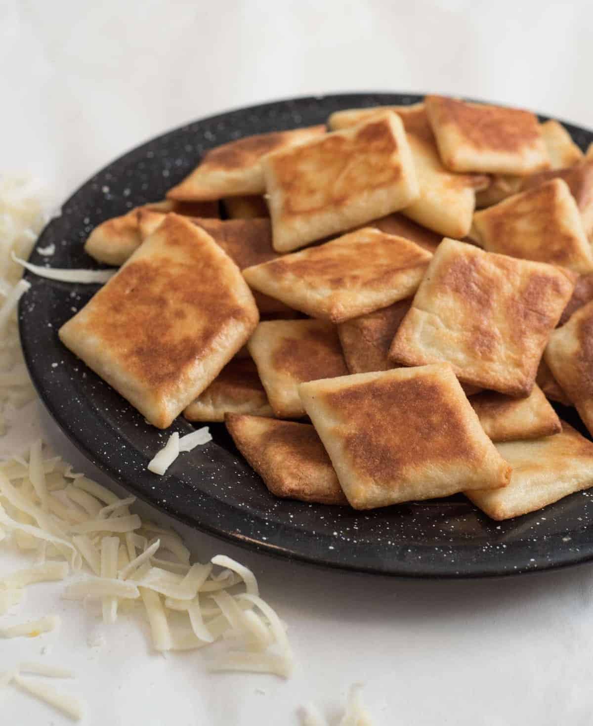 Close-up of baked fathead crackers on a black plate with white speckles on a white counter with some shredded mozzarella in the background. 