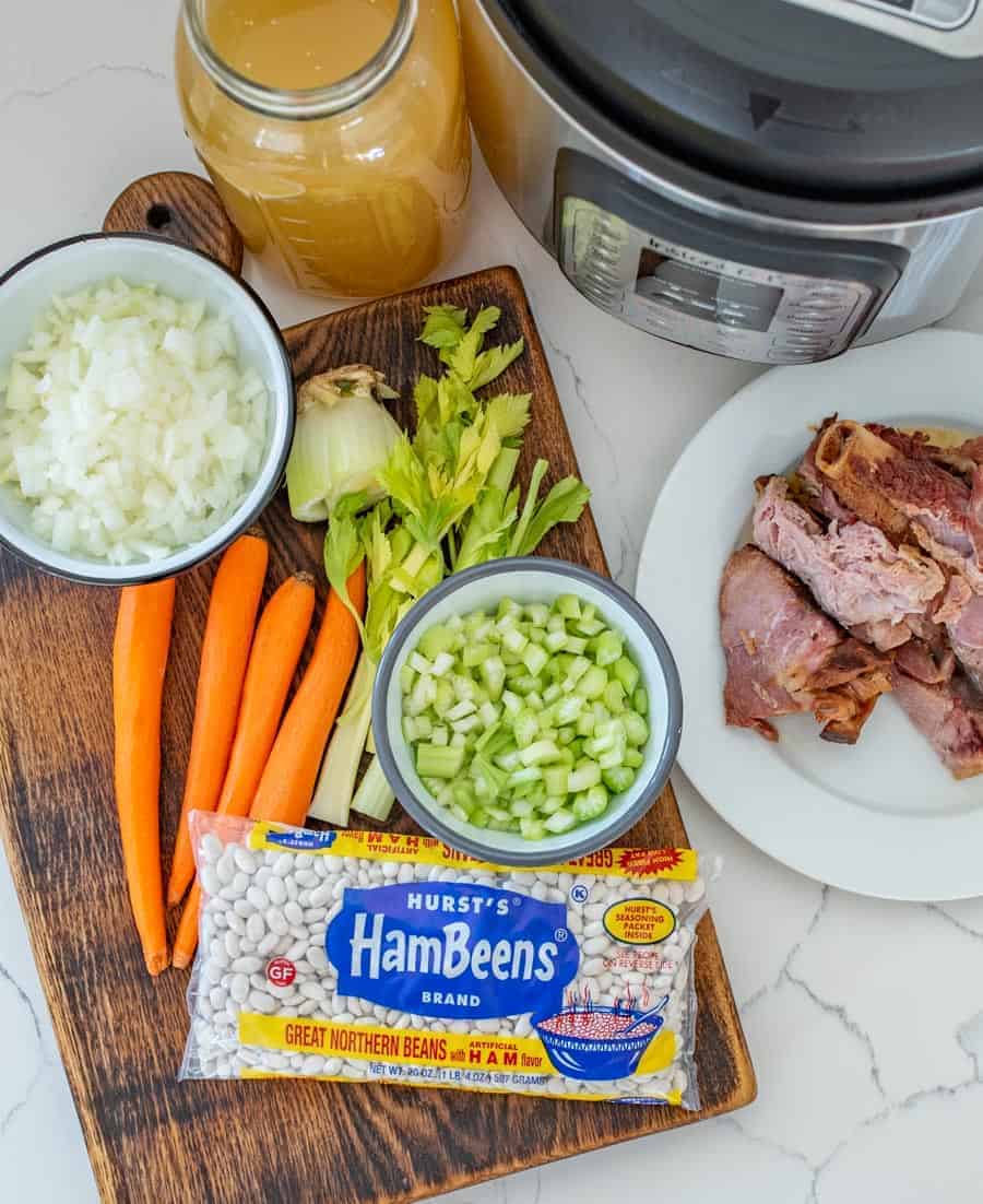 Image of all the ingredients including ham on a white plate, and hambeens great northern beans, carrots, celery, and onions on a wooden cutting board with an Instant Pot in the background. 