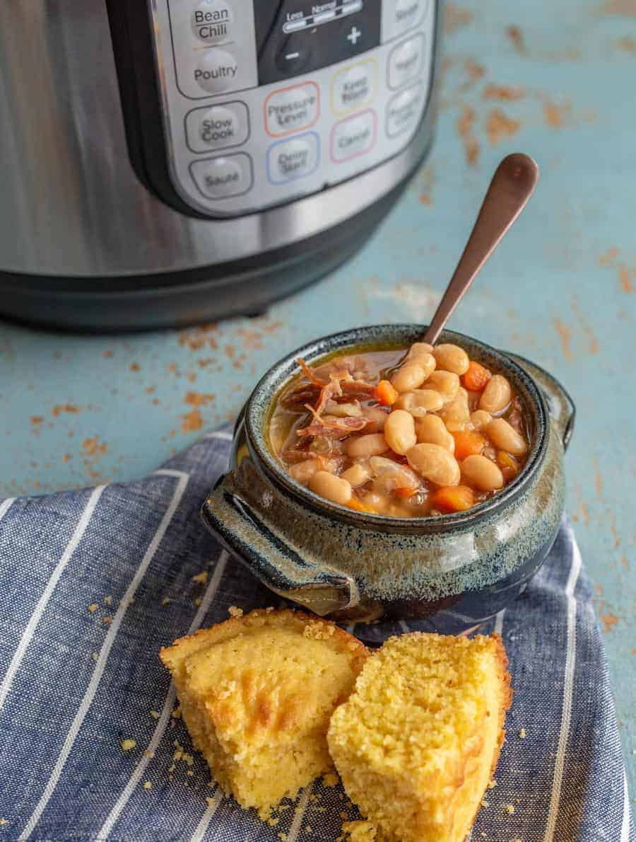 Image of a bowl of ham and bean soup in a blue pottery bowl with a soup spoon in it and a piece of cornbread in front of the bowl of soup on a blue napkin with white stripes with an Instant Pot in the background. 