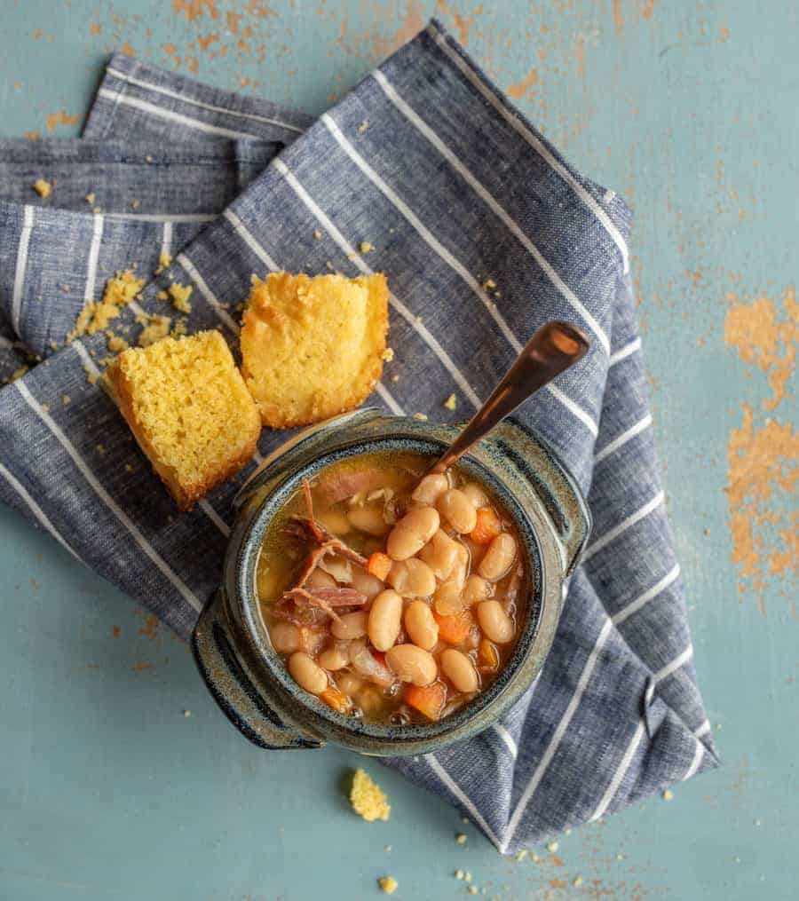 Image of a bowl of ham and bean soup in a blue pottery bowl with a soup spoon in it and a piece of cornbread in the background on a blue napkin with white stripes.