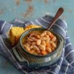 A rustic blue ceramic bowl brimming with a hearty ham and bean soup recipe, featuring succulent chunks of meat and diced carrots, is served with a spoon. The scene is completed by pieces of cornbread resting on a striped cloth atop the textured blue surface.
