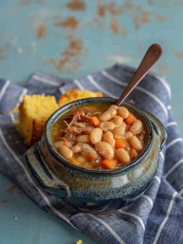 A rustic blue ceramic bowl brimming with a hearty ham and bean soup recipe, featuring succulent chunks of meat and diced carrots, is served with a spoon. The scene is completed by pieces of cornbread resting on a striped cloth atop the textured blue surface.
