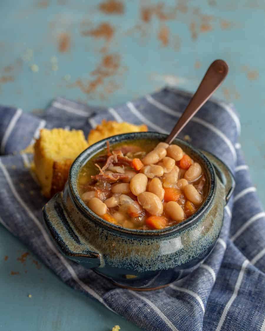 Image of a bowl of ham and bean soup in a blue pottery bowl with a soup spoon in it and a piece of cornbread in the background on a blue napkin with white stripes.