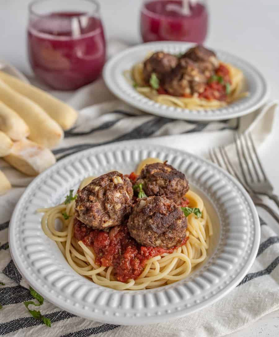 Homemade meatballs on top of a plate of spaghetti with marinara sauce with grape juice and bread sticks in the background. 