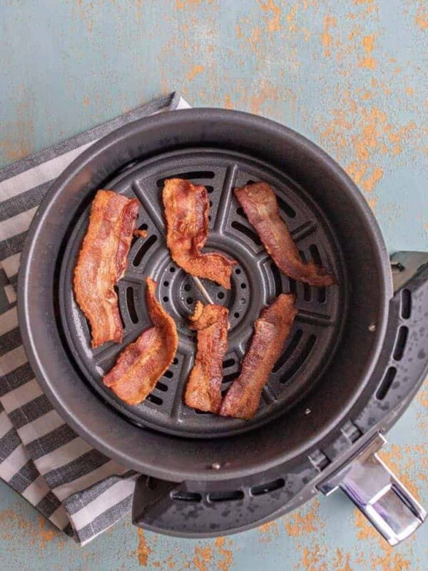 Crispy bacon in the Air Fryer, perfectly cooked strips rest in a basket atop a black and white striped napkin on a distressed blue surface.