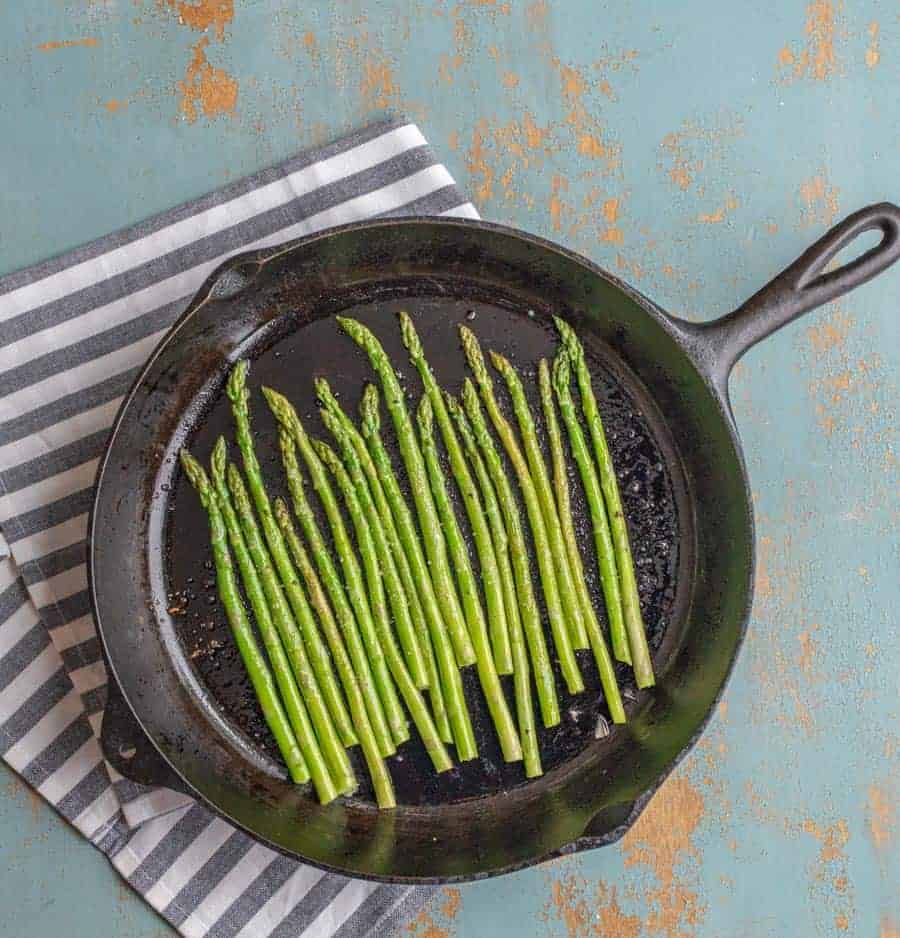 Bright green cooked asparagus left in whole spears in a line resting in a large black cast iron pan. 