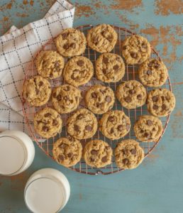 A cooling rack of peanut butter oatmeal and chocolate chip cookies rests on a rustic teal surface, accompanied by two glasses of milk. A checkered cloth is partially visible in the corner.
