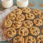 A batch of chocolate chip cookies cools on a round wire rack placed on a checkered cloth, accompanied by two glasses of milk. Nearby, a plate of peanut butter oatmeal cookies complements the rustic teal surface.
