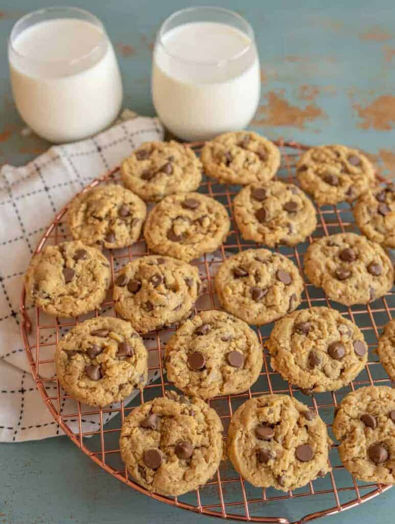 A batch of chocolate chip cookies cools on a round wire rack placed on a checkered cloth, accompanied by two glasses of milk. Nearby, a plate of peanut butter oatmeal cookies complements the rustic teal surface.