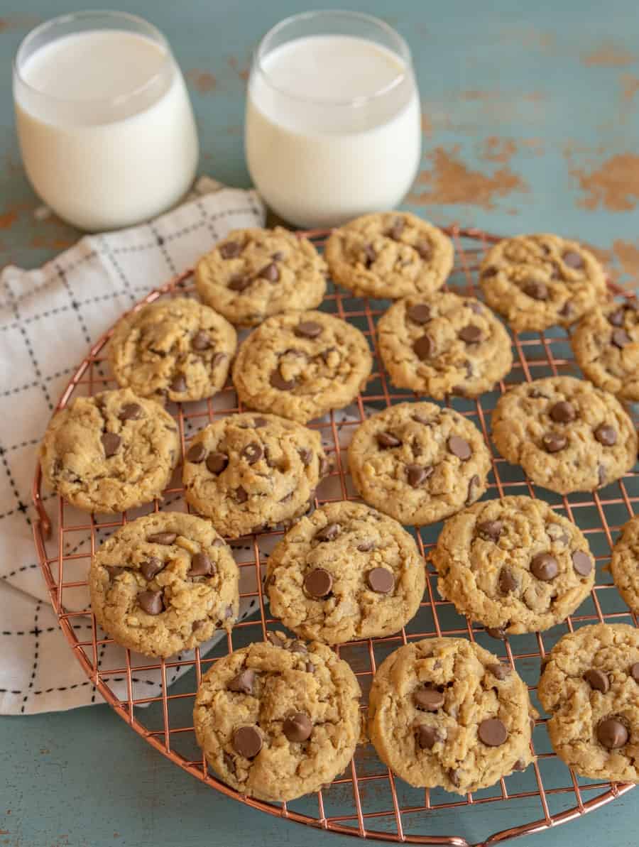 A batch of chocolate chip cookies cools on a round wire rack placed on a checkered cloth, accompanied by two glasses of milk. Nearby, a plate of peanut butter oatmeal cookies complements the rustic teal surface.