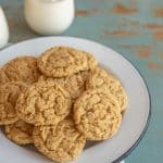 A white plate piled with peanut butter oatmeal cookies sits on a blue rustic table. In the background, two glasses of milk are visible, one with a cookie resting on top.