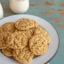 A white plate piled with peanut butter oatmeal cookies sits on a blue rustic table. In the background, two glasses of milk are visible, one with a cookie resting on top.